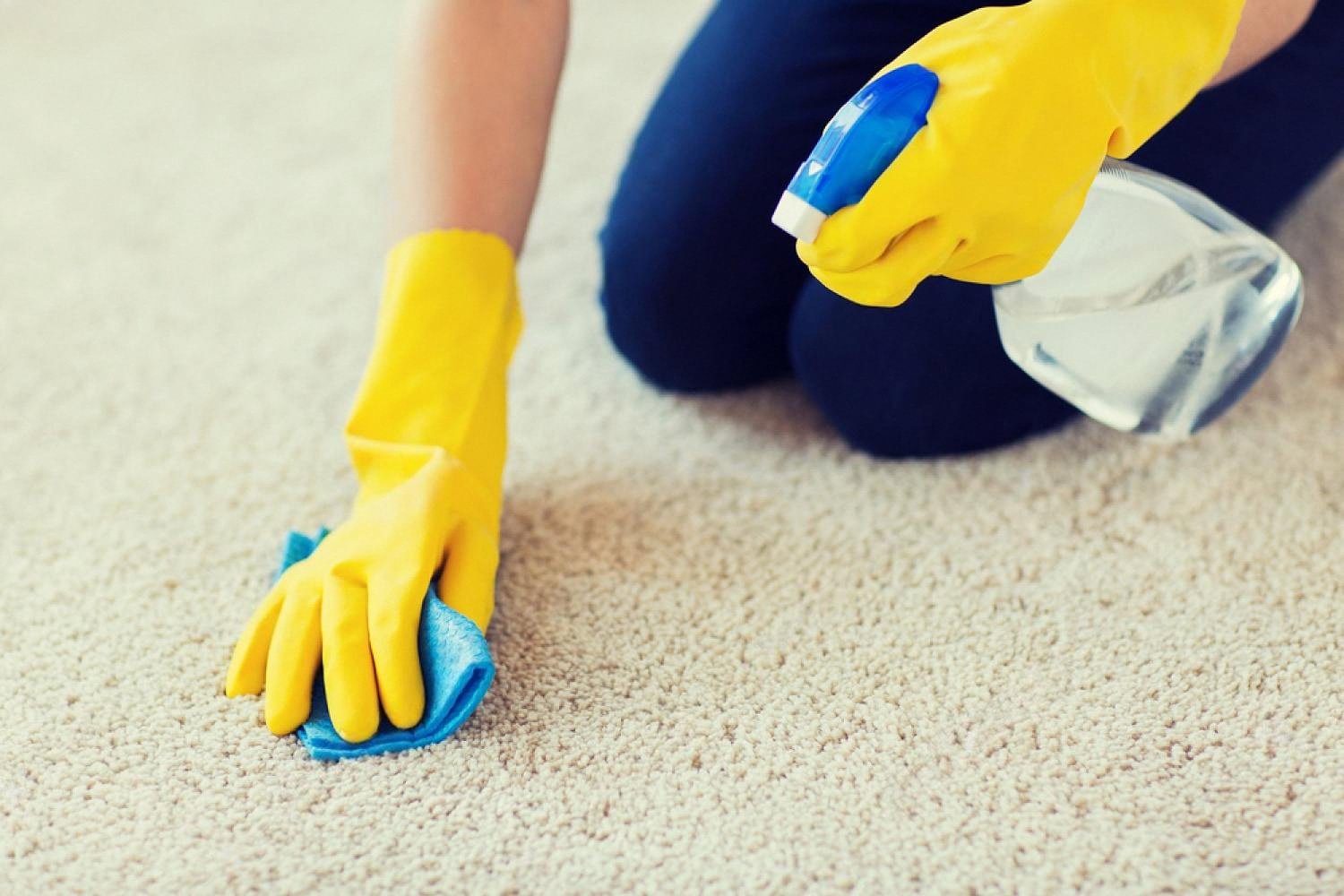 Person in yellow gloves spraying and wiping a beige carpet with a cloth